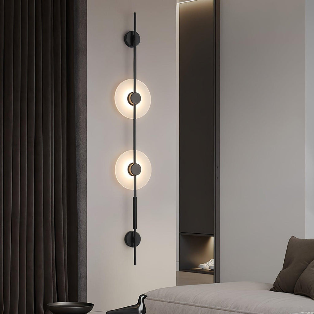 Black Clérachic vertical sconce with two lit alabaster discs on a pale wall beside dark curtains, with a sofa edge at the lower right.