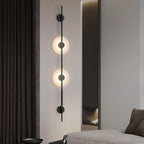 Black Clérachic vertical sconce with two lit alabaster discs on a pale wall beside dark curtains, with a sofa edge at the lower right.
