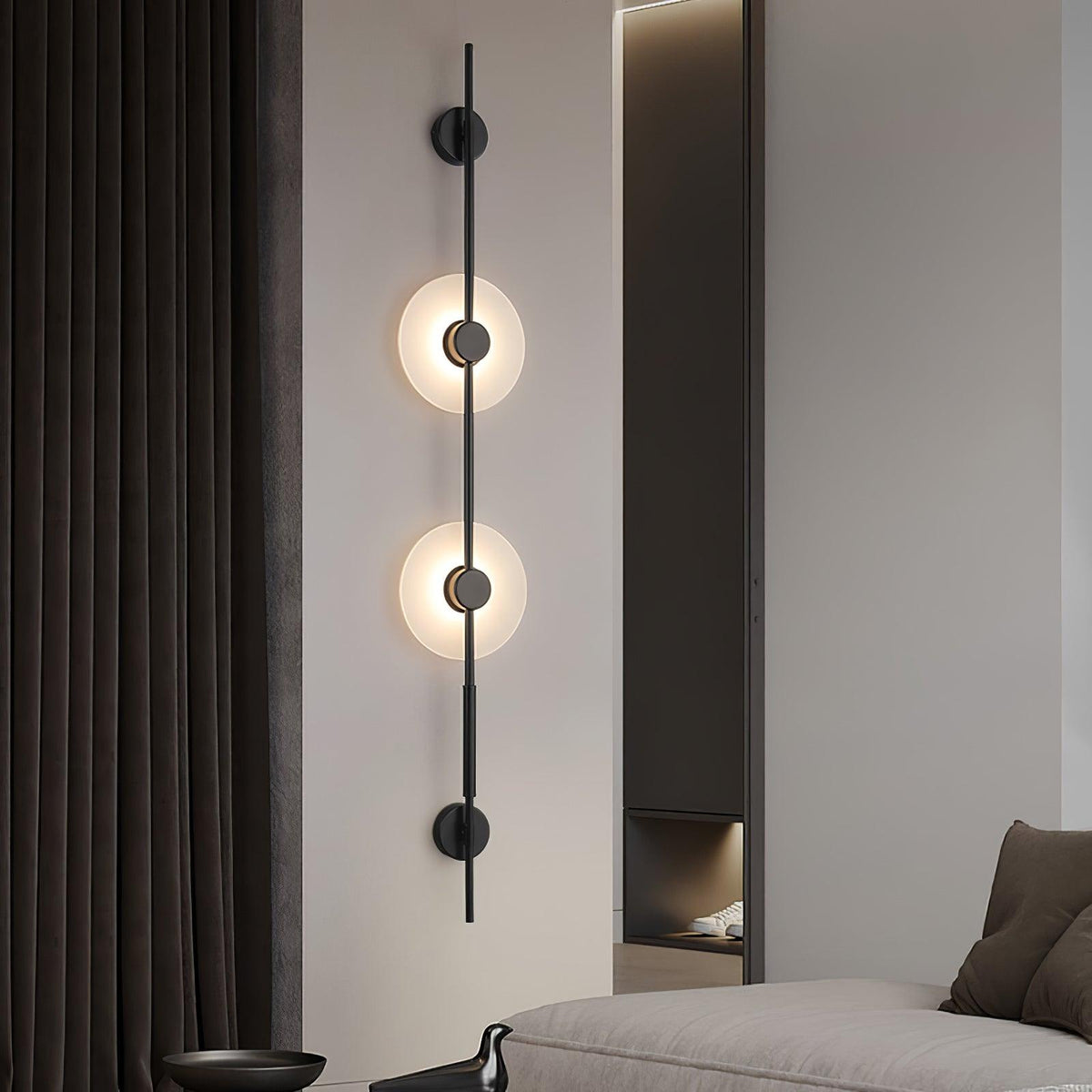 Black Clérachic vertical sconce with two lit alabaster discs on a pale wall beside dark curtains, with a sofa edge at the lower right.