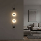 Black Clérachic vertical sconce with two glowing alabaster discs on a grey wall, with a pale sofa and a framed line drawing beyond.