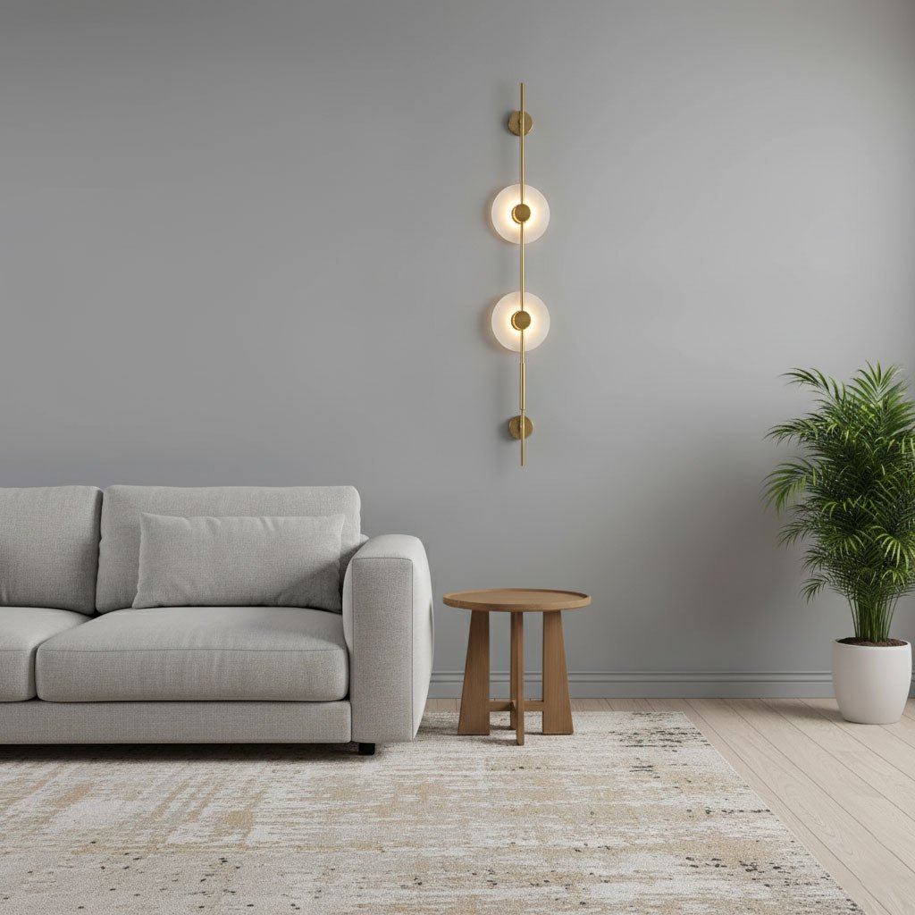 Gold Clérachic sconce with two lit alabaster discs on a pale grey wall, beside a light sofa, a round wooden table and a potted palm.
