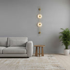 Gold Clérachic sconce with two lit alabaster discs on a pale grey wall, beside a light sofa, a round wooden table and a potted palm.