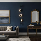 Three-disc black Clérachic sconce lit on a navy wall beside a gilt-framed mirror, with a grey sofa and a brass-legged console.
