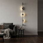 Black Clérachic sconce with three softly lit discs on a pale wall, above a dark armchair, a side table and a draped throw.