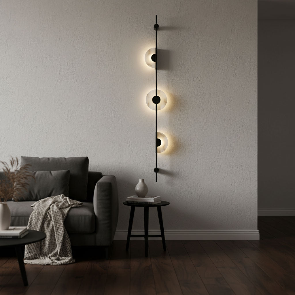 Black Clérachic sconce with three softly lit discs on a pale wall, above a dark armchair, a side table and a draped throw.