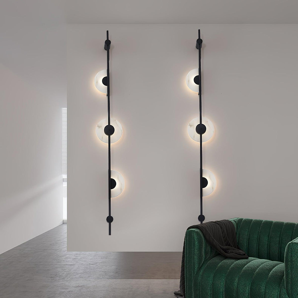 Two black three-disc Clérachic sconces lit on a white wall, with the arm of a green velvet sofa at the lower right.
