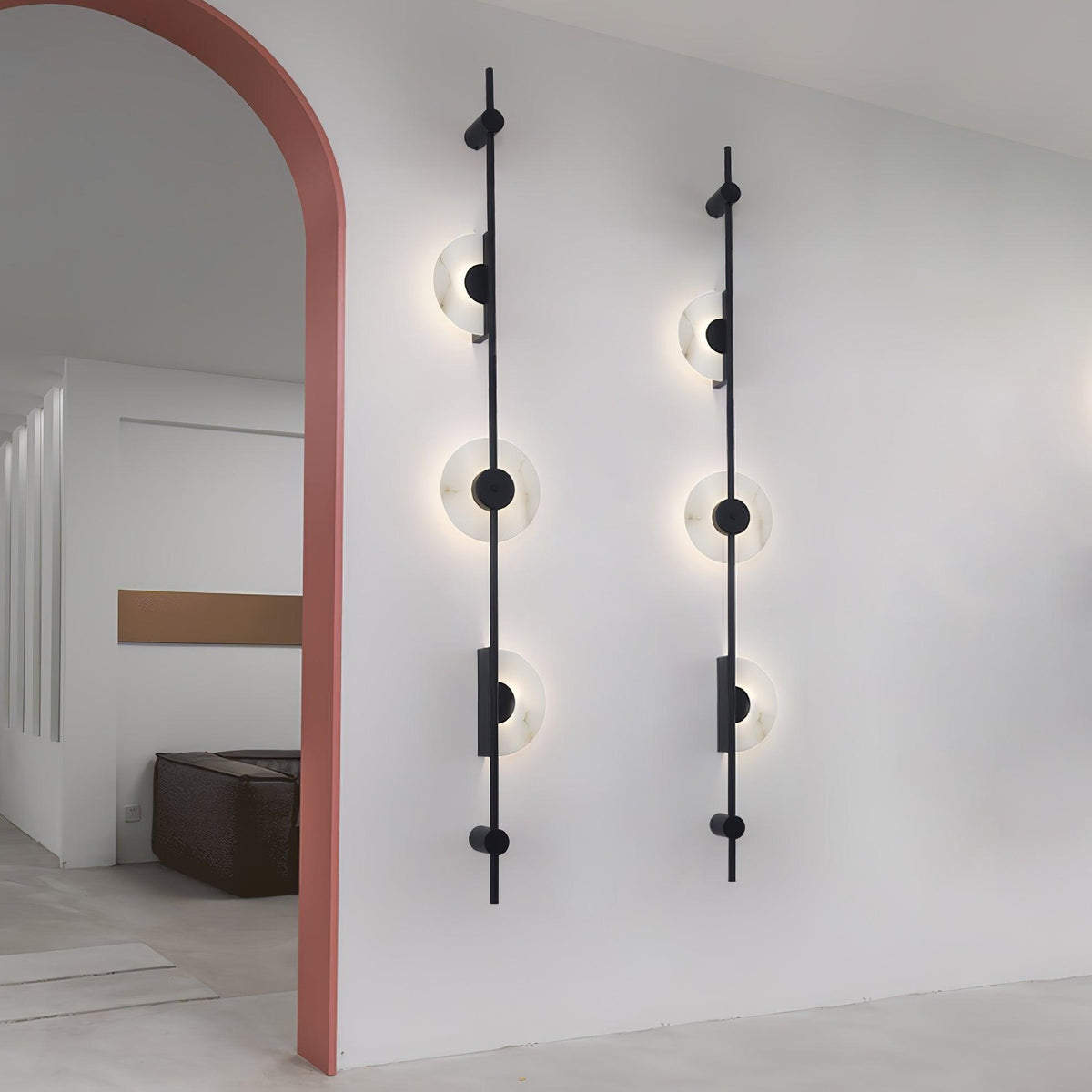 A pair of black three-disc Clérachic sconces on a white wall beside a terracotta-pink arch, with a lit room beyond.