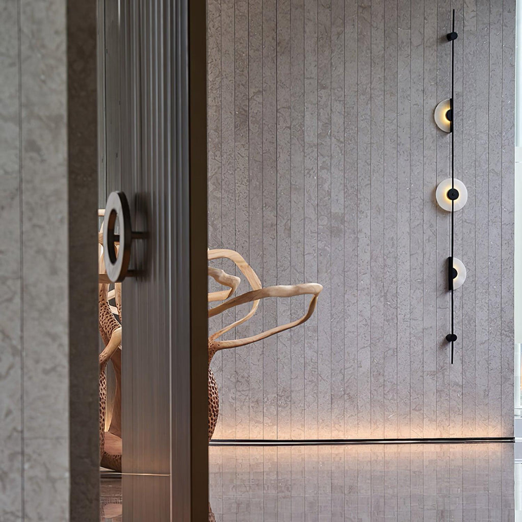 Black Clérachic sconce running up a textured grey wall behind a fluted column, with its three discs lit and a wood sculpture below.