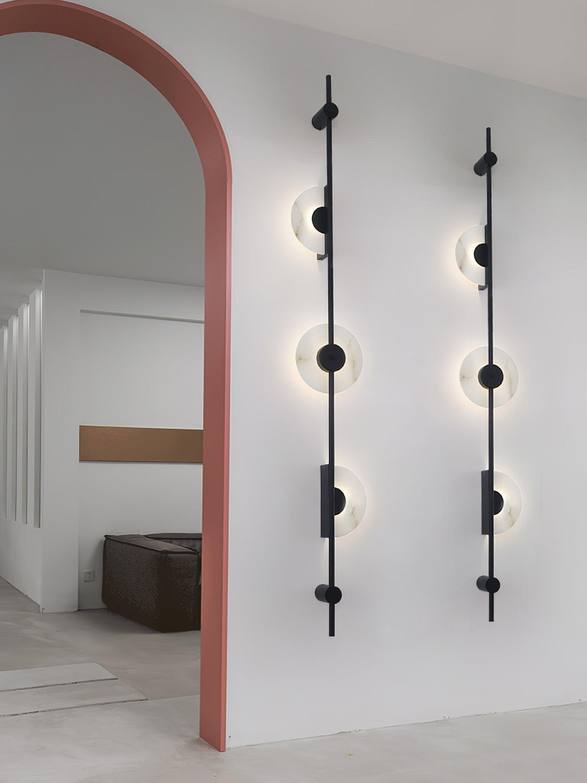 Two black three-disc Clérachic sconces side by side on a white wall beside a pink arch, with the discs glowing.