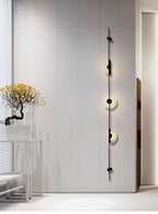 Distant view of a black three-disc Clérachic sconce on a tall white wall, beside a small tree on a console.
