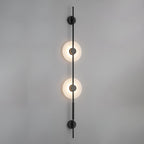 Close front view of this Clérachic sconce in black, two lit alabaster discs on a rod against a plain grey wall.