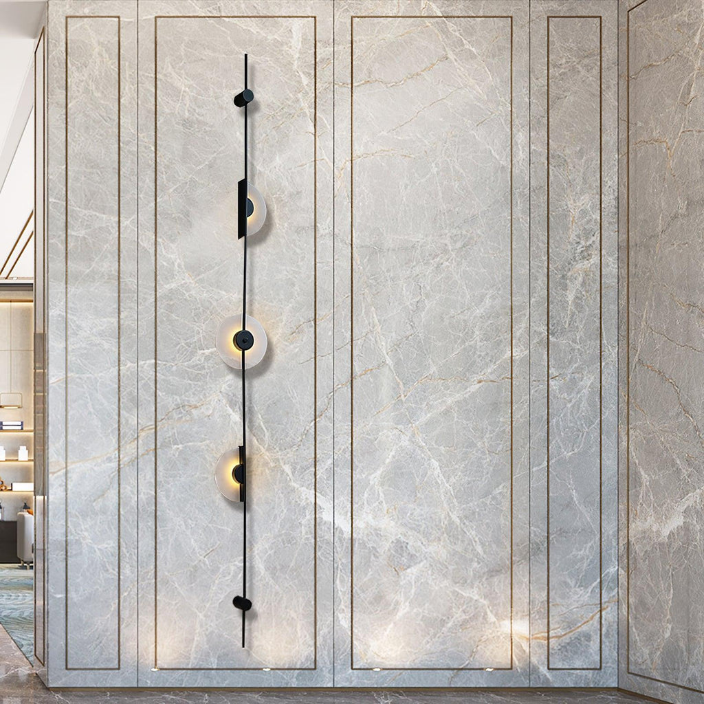 Black three-disc Clérachic sconce on a marble-look panelled wall, its discs lit and a warm glow near the floor.
