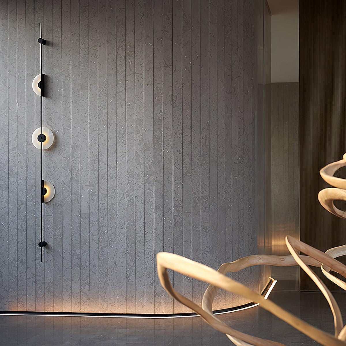 Black Clérachic sconce on a grey slatted wall, lit, with a sweeping pale wood sculpture and a floor strip light below.