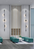 A pair of black three-disc Clérachic sconces on a tall pale wall flanking slatted wood panelling, above teal armchairs in a lobby.