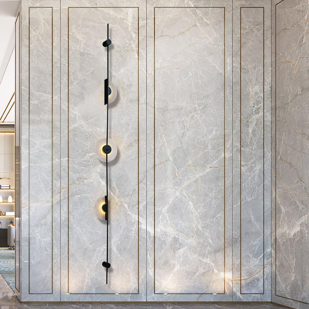Tall black three-disc Clérachic sconce on a grey marble-look panel with fine gold veining, its discs glowing.