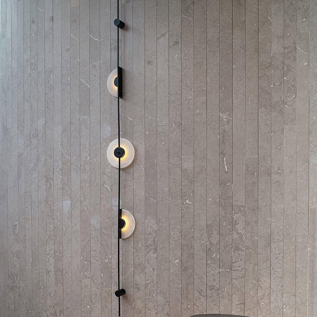 Close view of a black Clérachic sconce on a grey slatted stone wall, three alabaster discs lit along the rod.