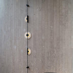 Close view of a black Clérachic sconce on a grey slatted stone wall, three alabaster discs lit along the rod.