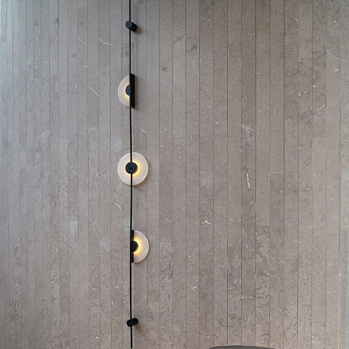 Close view of a black Clérachic sconce on a grey slatted stone wall, three alabaster discs lit along the rod.
