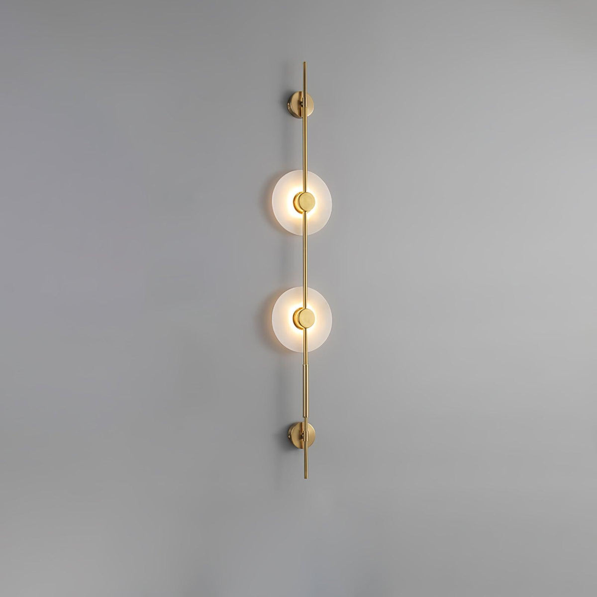 Gold Clérachic sconce with two glowing alabaster discs on a plain grey wall, a slim rod running above and below.