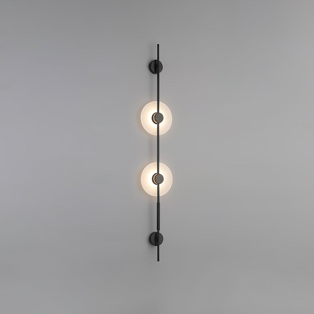 Black Clérachic sconce with two lit alabaster discs on a plain grey wall, with round black caps at both ends.