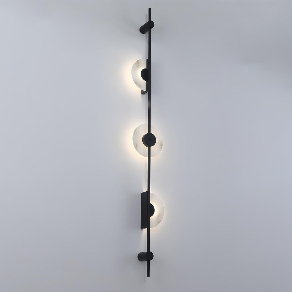 Black three-disc Clérachic sconce on a pale wall seen at an angle, each half-disc glowing around the rod.