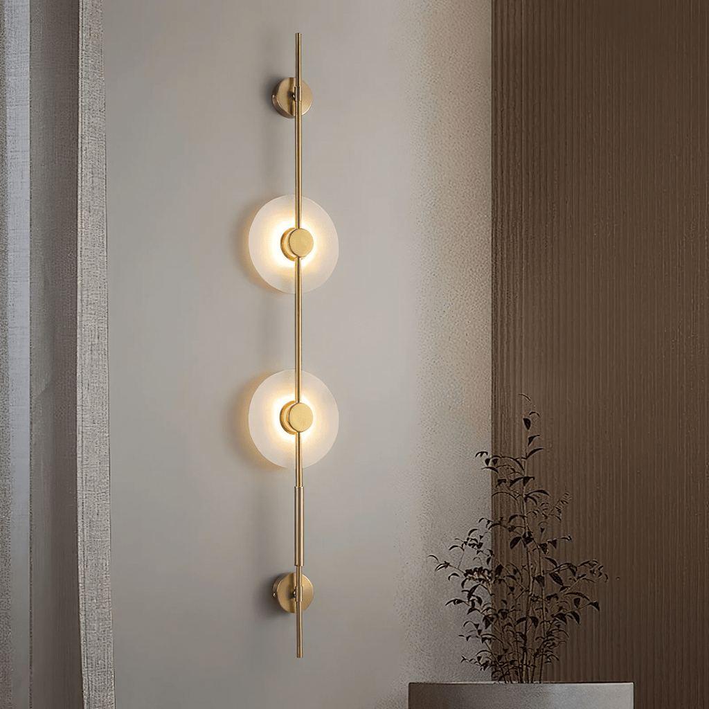 Gold Clérachic sconce with two lit alabaster discs on a warm wall beside wood panelling, with dried branches in a pot below.