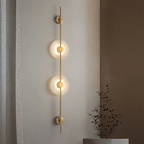 Gold Clérachic sconce with two lit alabaster discs on a warm wall beside wood panelling, with dried branches in a pot below.