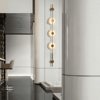 Gold three-disc Clérachic sconce lit on a white column in a bright interior, with a bar stool at the left.