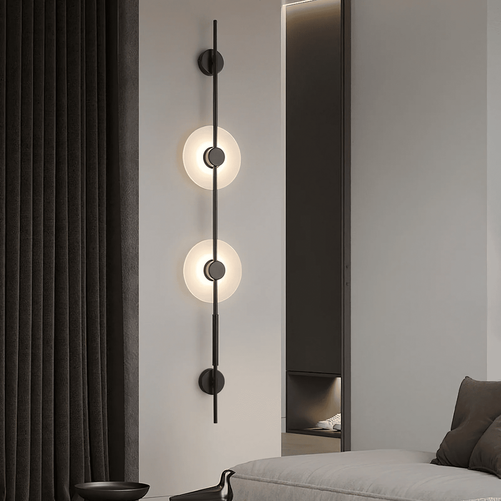 Black Clérachic sconce with two lit alabaster discs on a pale wall, beside dark curtains and a sofa at the bottom.