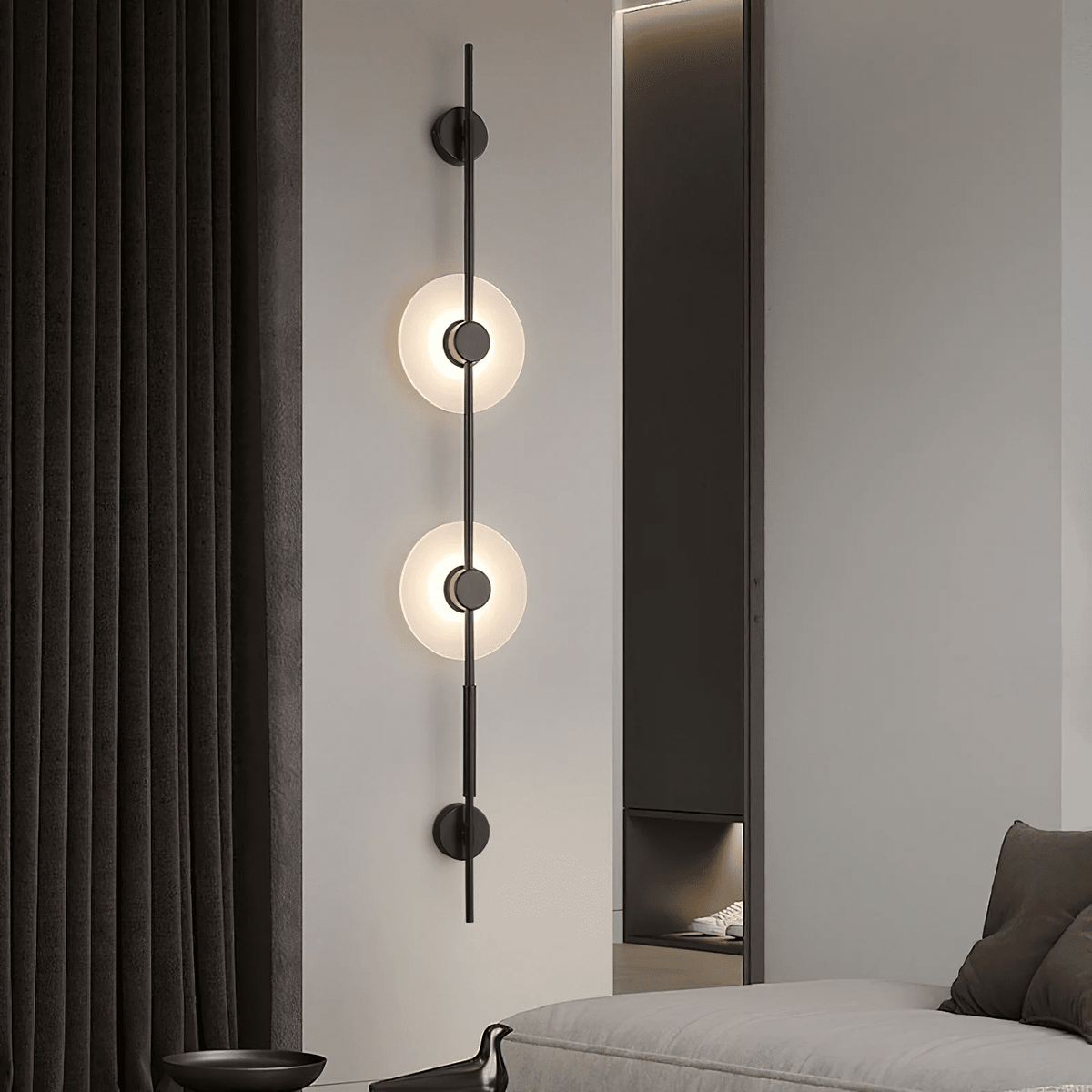 Black Clérachic sconce with two lit alabaster discs on a pale wall, beside dark curtains and a sofa at the bottom.