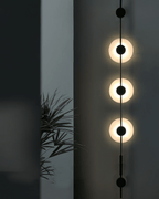 Close view of a black Clérachic sconce with three brightly glowing alabaster discs, a dark wall and a palm leaf behind.