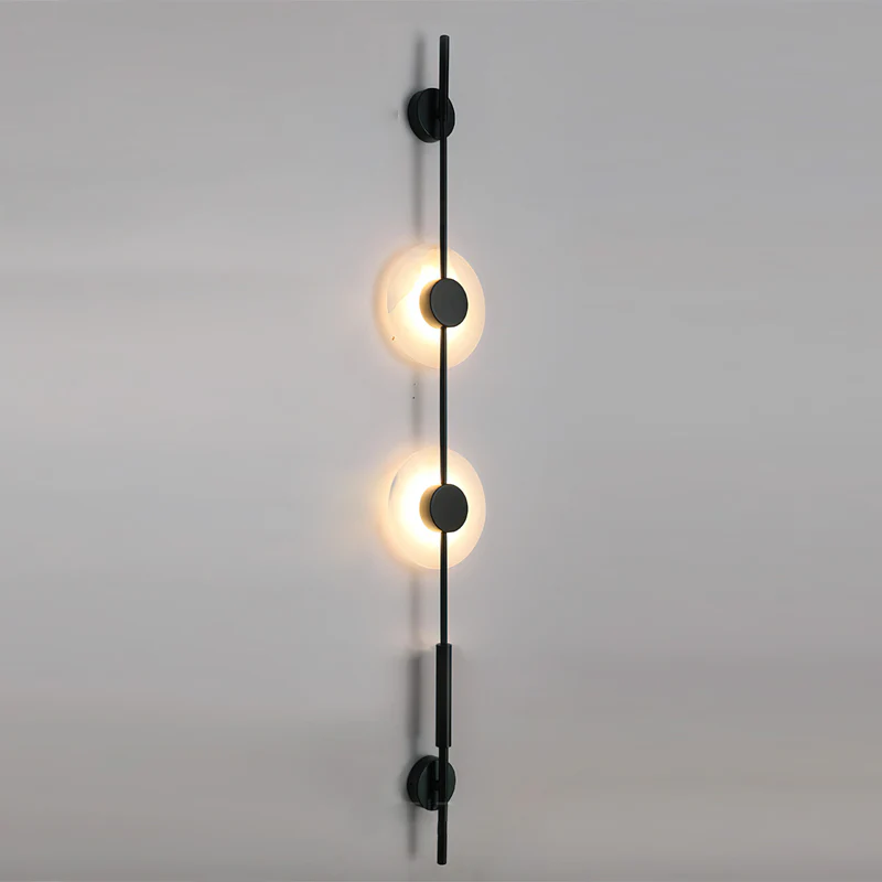 Black Clérachic sconce with two warmly lit alabaster discs on a grey wall, seen at a slight angle from below.