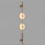 Gold Clérachic sconce with two lit alabaster discs on a grey wall, with a long rod and brass wall caps at both ends.