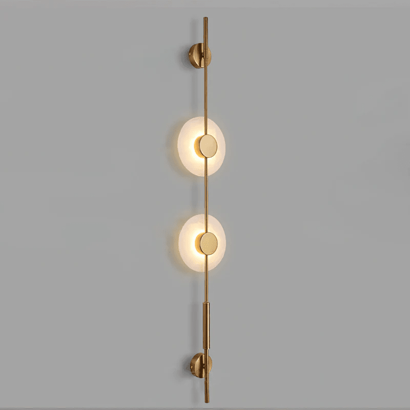 Gold Clérachic sconce with two lit alabaster discs on a grey wall, with a long rod and brass wall caps at both ends.