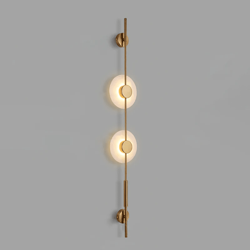 Gold Clérachic sconce with two lit alabaster discs on a grey wall, with small round brass caps at the top and bottom.