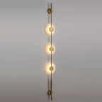 Gold three-disc Clérachic sconce with warmly lit alabaster discs on a grey wall, seen from a slight angle.
