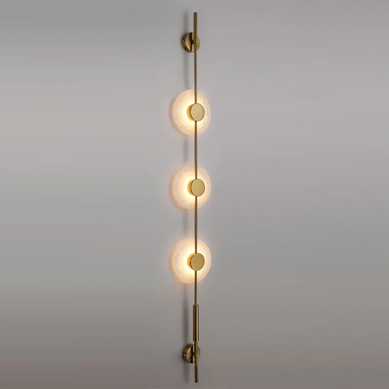 Gold three-disc Clérachic sconce with warmly lit alabaster discs on a grey wall, seen from a slight angle.