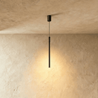 A slim Clérachic black pendant rod drops from a textured ceiling mount, its lower tip glowing warm against plaster.