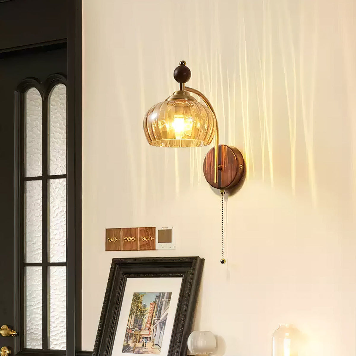 Lit amber glass Clérachic wall light with a walnut backplate and pull chain, beside a framed print and a dark-framed window.