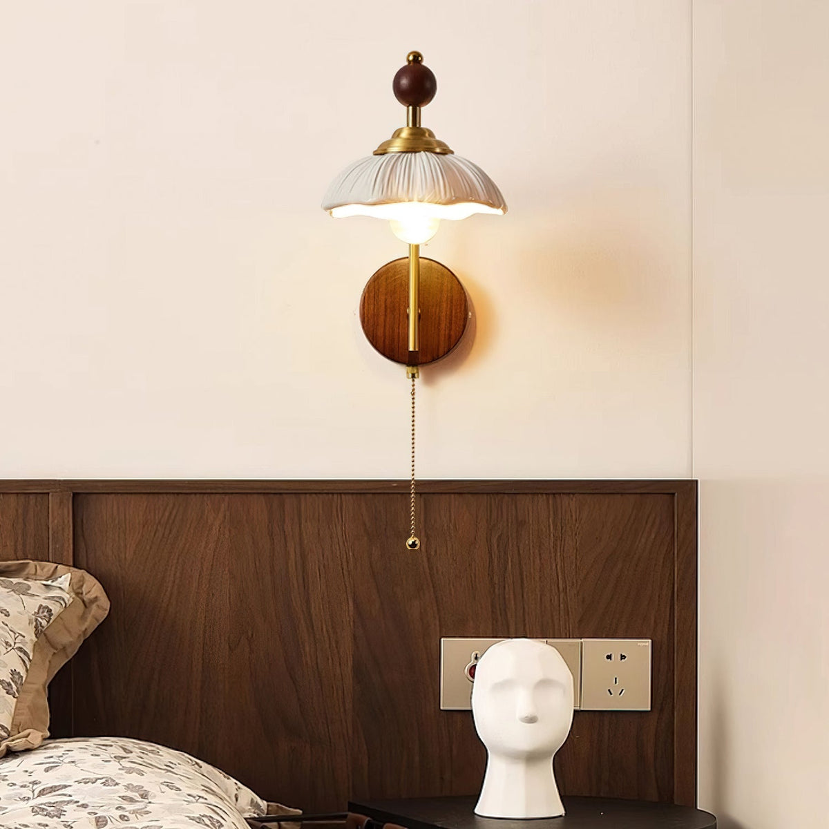 Clérachic wall light with a scalloped white shade and pull chain, above a walnut headboard and a pale head-shaped ornament.