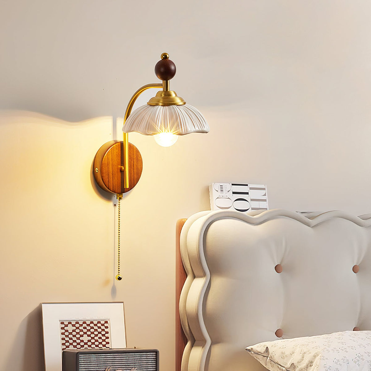 A Clérachic wall light with a white scalloped shade lit above a cream scalloped headboard and a framed print.