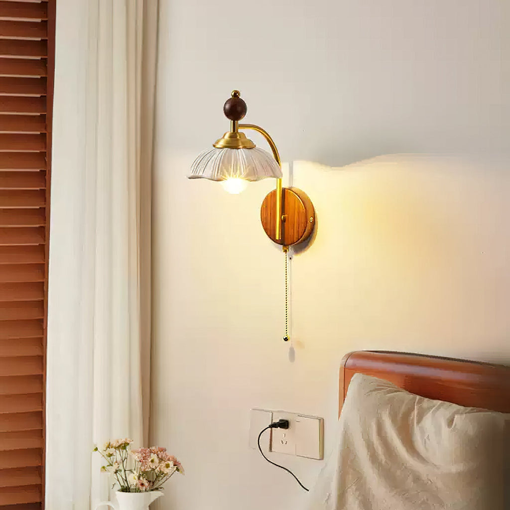 A Clérachic wall light with a white scalloped shade and pull chain, beside a louvred shutter, a curtain and a wooden bedhead.