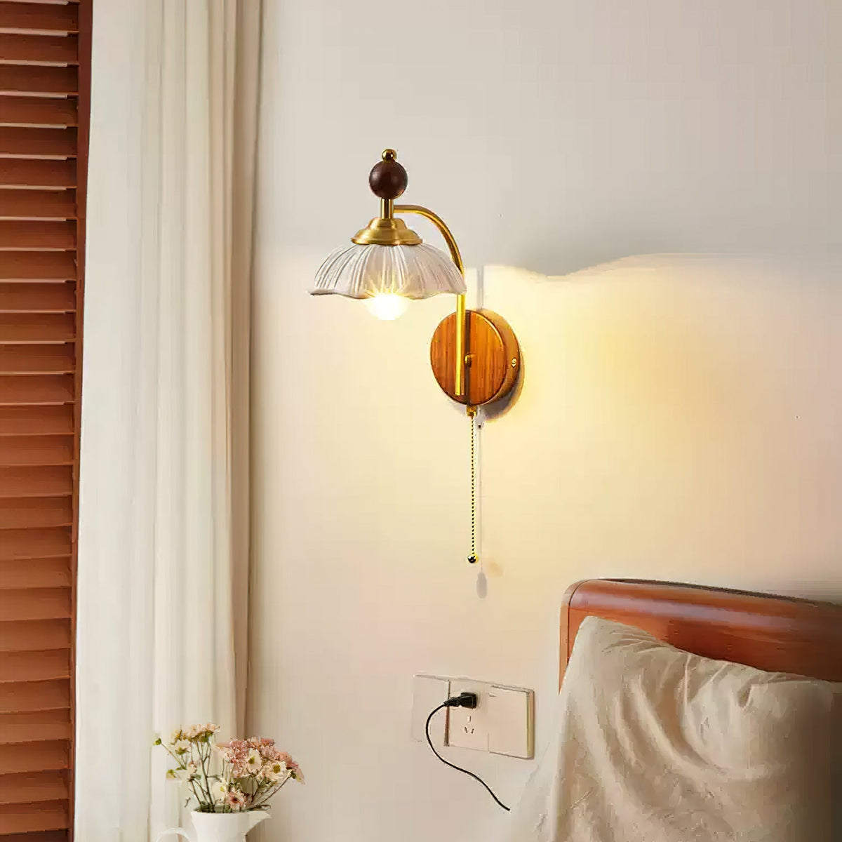 A Clérachic wall light with a white scalloped shade and pull chain, beside a louvred shutter, a curtain and a wooden bedhead.