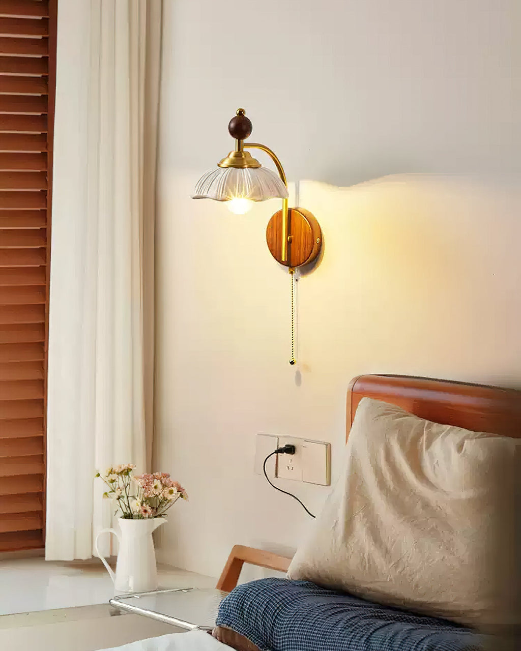 Clérachic wall light with a white scalloped shade lit beside a curtain, above a bed with a wooden frame and a jug of flowers.