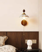 White scalloped-shade Clérachic wall light on a cream wall, above a walnut headboard and a dark bedside table.