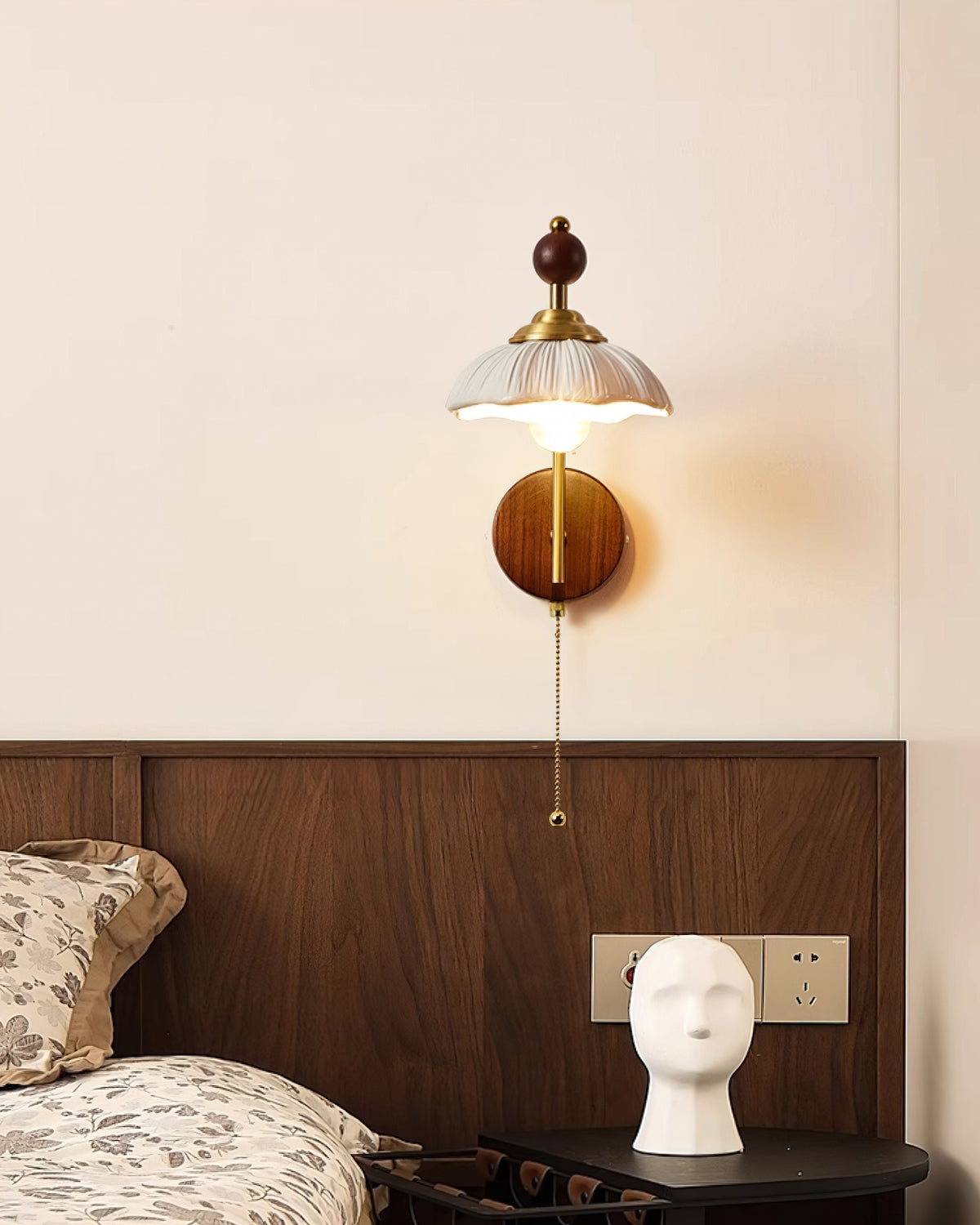 White scalloped-shade Clérachic wall light on a cream wall, above a walnut headboard and a dark bedside table.