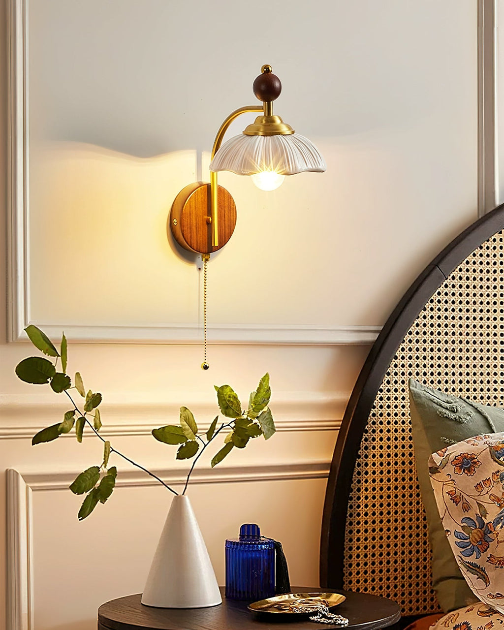 Lit Clérachic wall light with a white scalloped shade beside a rattan bedhead, above a bedside table with a leafy stem in a vase.