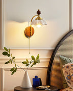 Lit Clérachic wall light with a white scalloped shade beside a rattan bedhead, above a bedside table with a leafy stem in a vase.
