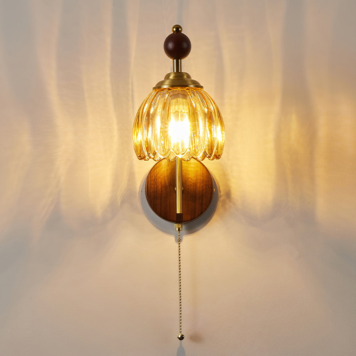 A lit amber glass Clérachic wall light with a walnut backplate and pull chain, throwing warm rippled light on the wall.