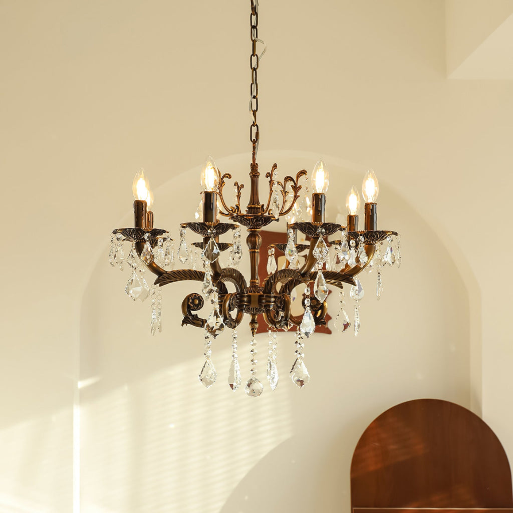 A Clérachic chandelier with bronze scrolled arms and clear prisms hangs from a chain before an arched cream wall, bulbs lit.